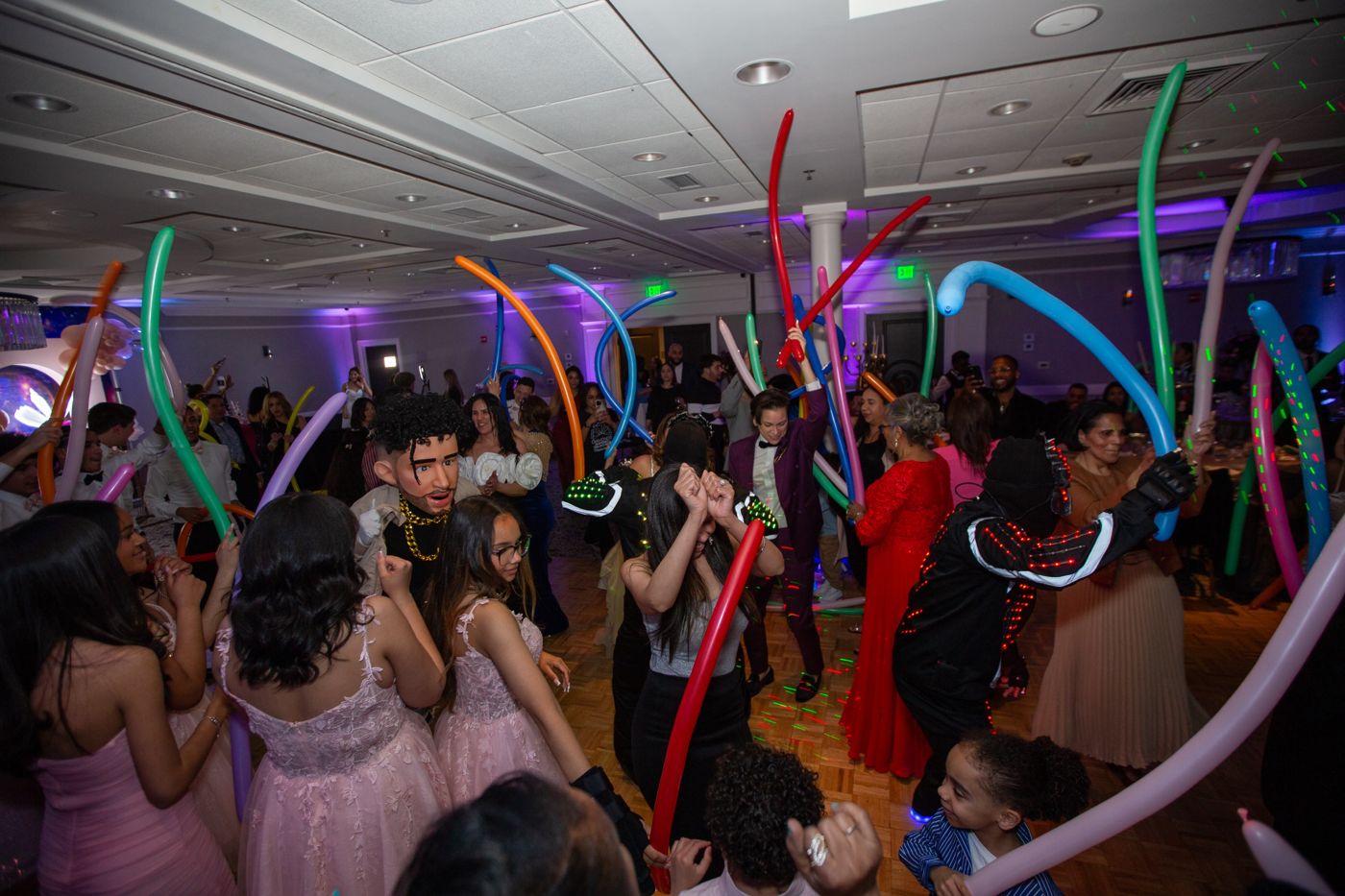Guests, costumed performers, and long balloons filling a quinceañera dance floor