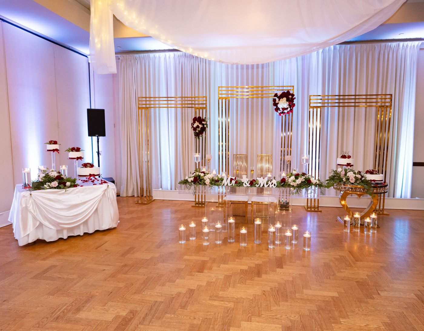 A sweetheart table with gold arches, candles, flowers, and cakes under soft uplighting